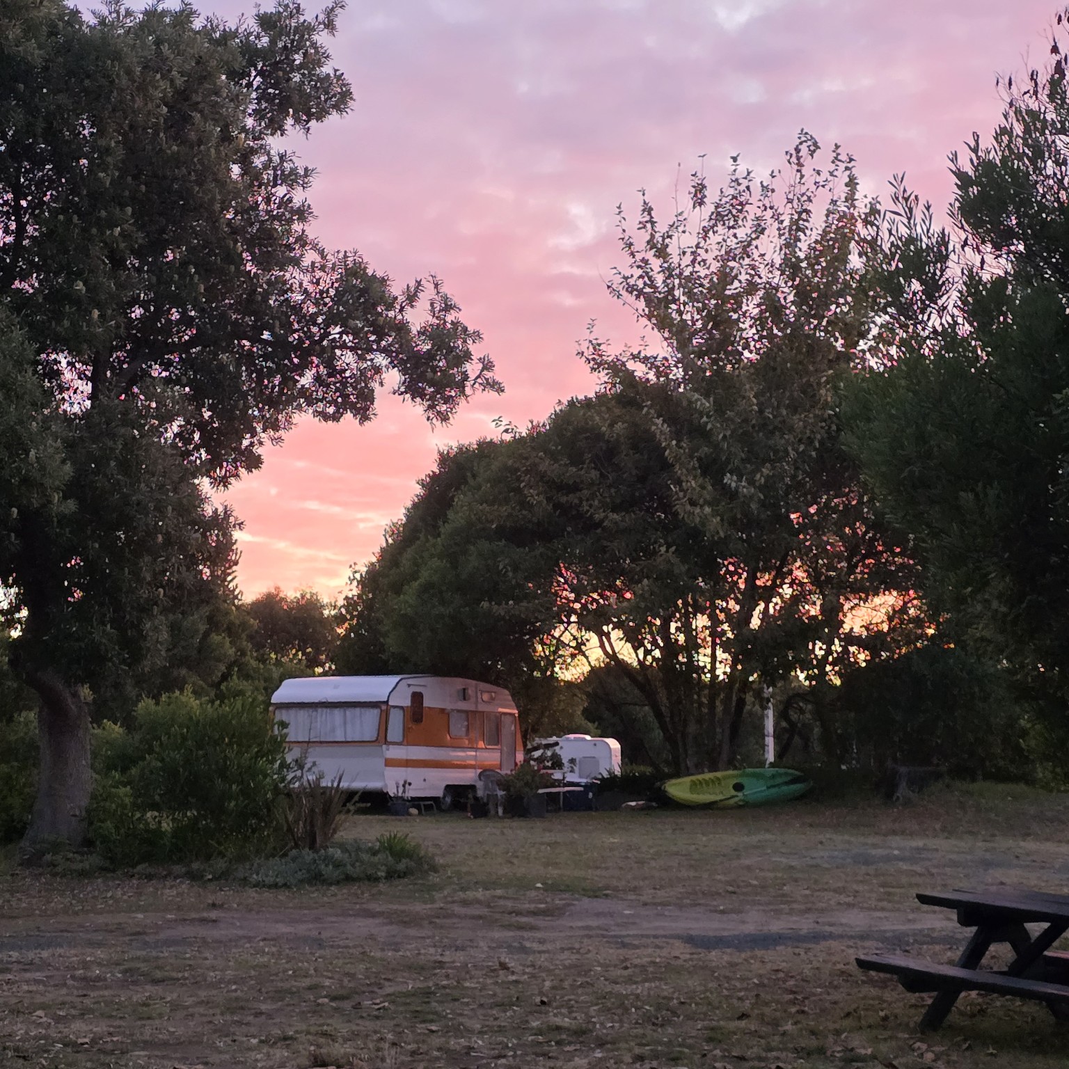 A vintage caravan parked under trees beneath a pink and orange evening sky