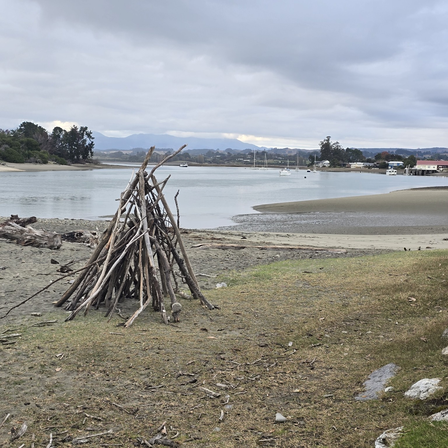 A driftwood shelter standing on the sand of a wide grey estuary
